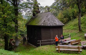 Eine kleine, h&ouml;lzerne Kirche im Wald mit zwei Personen und Holzb&auml;nken im Vordergrund.