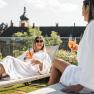 Zwei Frauen in Badem&auml;nteln entspannen auf Liegest&uuml;hlen mit Getr&auml;nken vor einem Schloss.