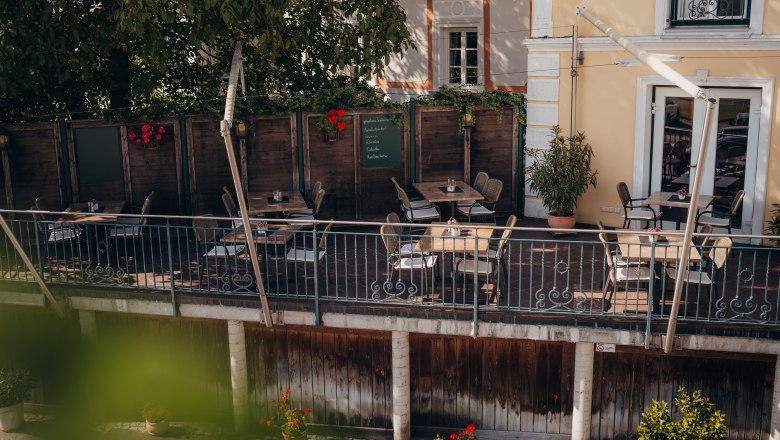 Gemütliche Terrasse eines Cafés mit Holztischen und Stühlen, umgeben von Pflanzen.