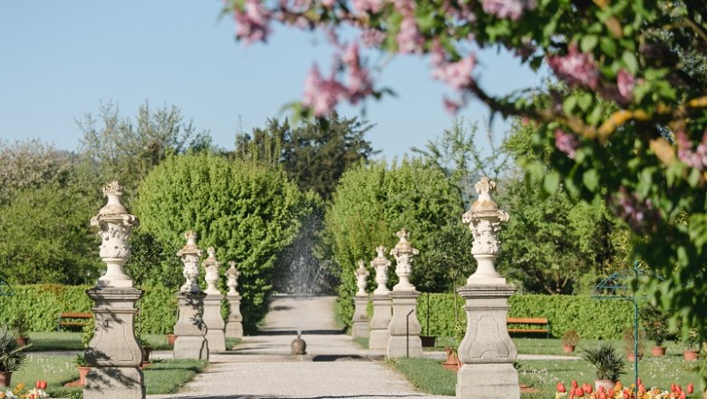Ein gepflegter Gartenweg mit Statuen und blühenden Bäumen im Frühling.