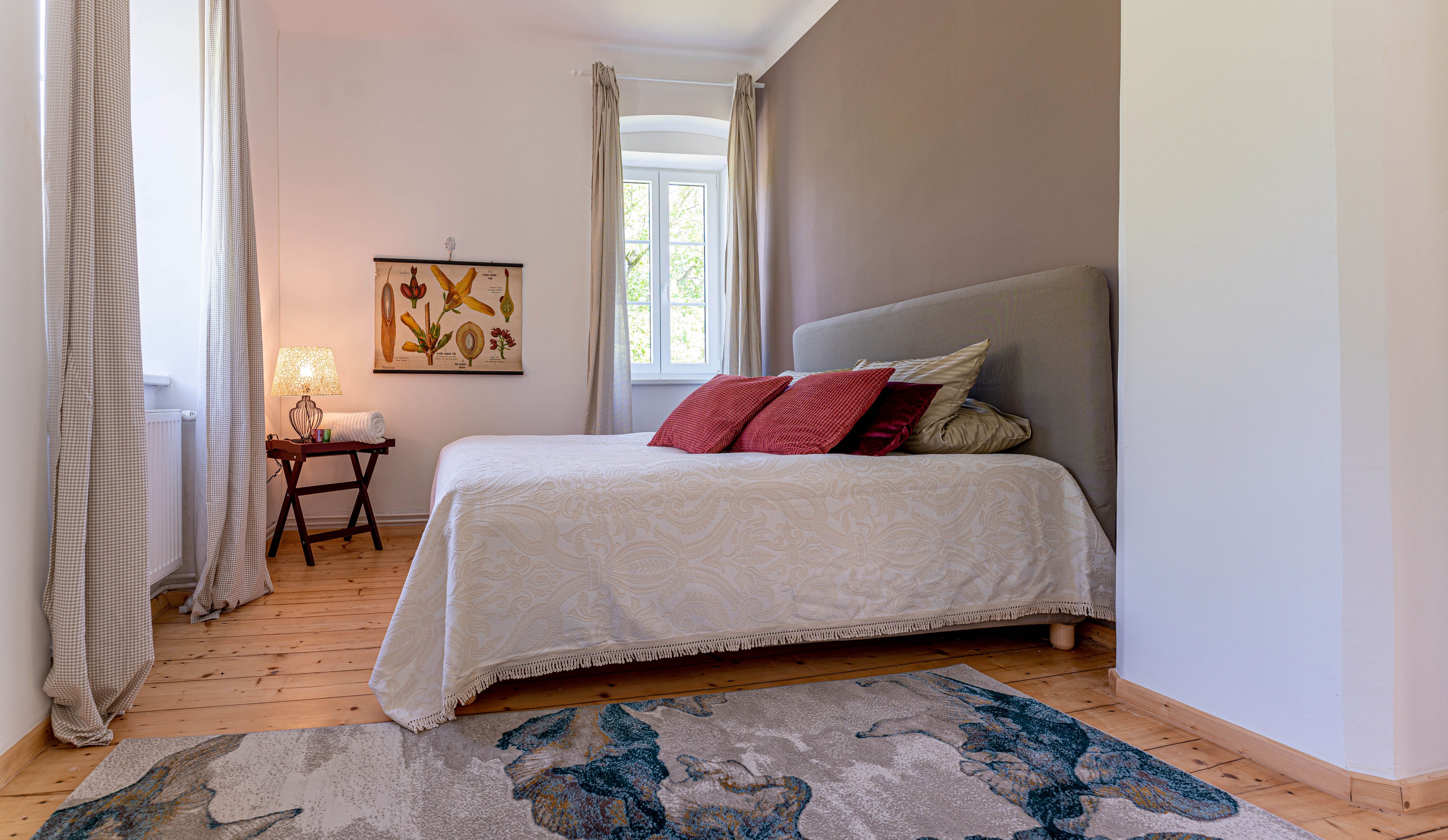 Schlafzimmer mit Bett, roten Kissen, Teppich und Fenster mit Vorhängen.