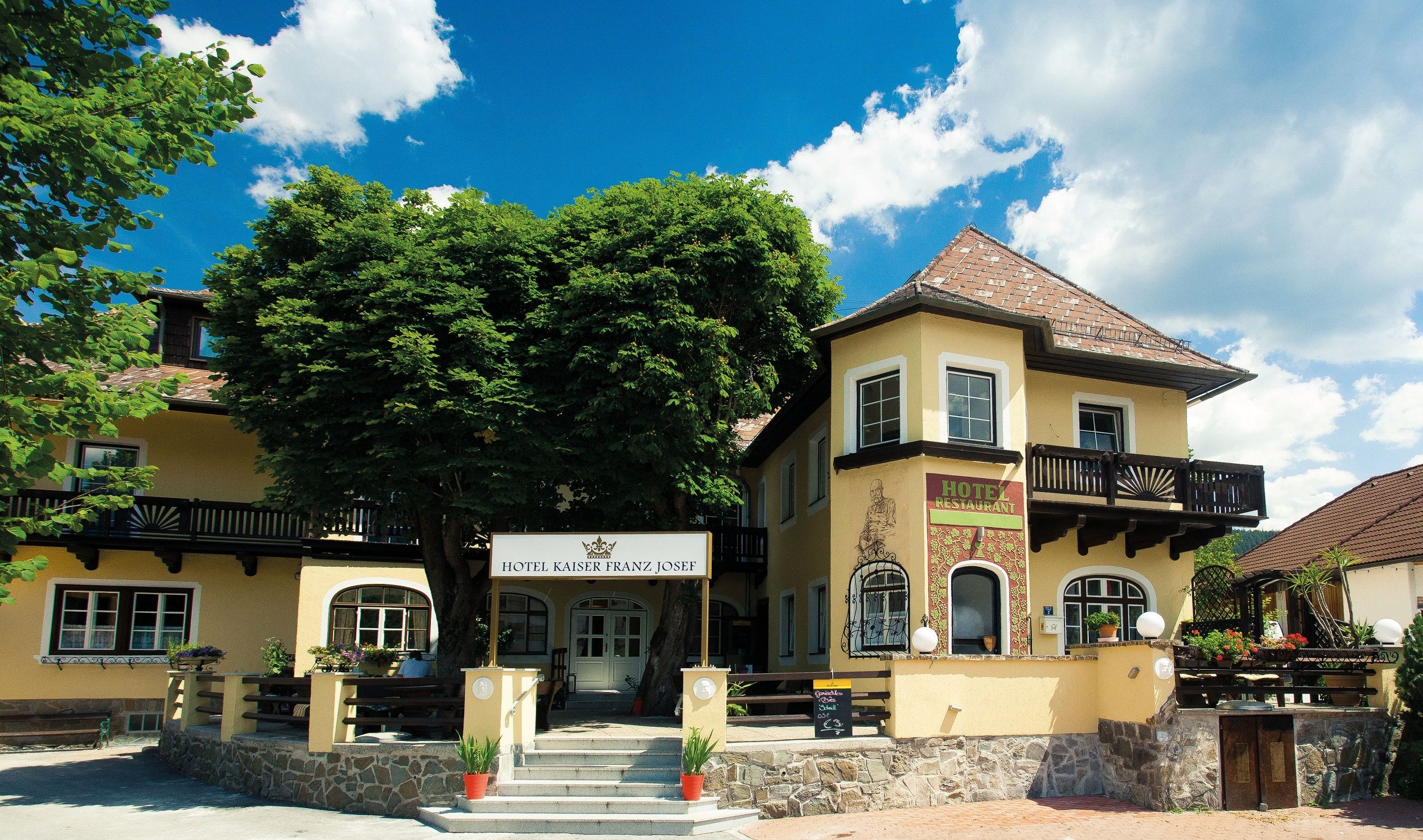 Außenansicht des Hotels Kaiser Franz Josef mit gelber Fassade und Bäumen.