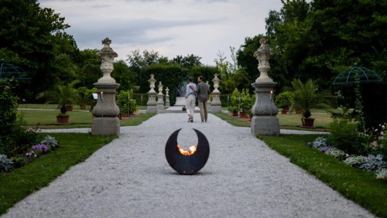 Ein Kiesweg im historischen Hofgarten des Stifts Seitenstetten mit Skulpturen und Pflanzen, im Vordergrund eine Feuerstelle.