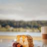 Kuchenstück mit Erdbeeren und Latte Macchiato vor unscharfer Landschaft.