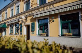 Au&szlig;enansicht eines gelben Geb&auml;udes mit der Aufschrift 'Zum goldenen L&ouml;wen'. Fenster mit Blumenk&auml;sten und gr&uuml;nen Markisen.