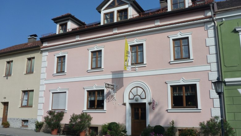 Au&szlig;enansicht eines rosa Hauses mit wei&szlig;en Fensterrahmen und einer gelben Fahne.
