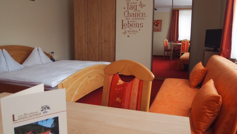 Ein gemütliches Hotelzimmer mit einem Doppelbett, einem Sofa, einem Tisch und einem Schrank. An der Wand steht ein motivierender Spruch.