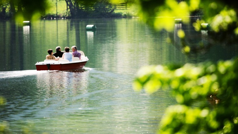 Vier Personen in einem Boot auf einem ruhigen See, umgeben von gr&uuml;ner Natur.