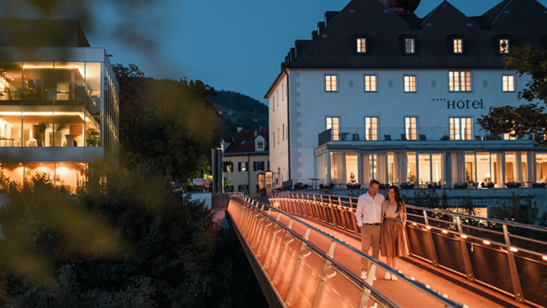 Ein Paar spaziert abends über eine beleuchtete Brücke vor einem Hotel.
