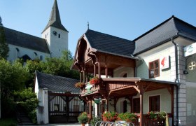 Ein traditionelles Gebäude mit Holzbalkon und Blumendekoration, daneben eine Kirche mit Turm.