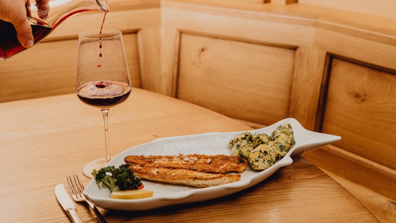 Zander in Knoblauchbutter, © Niederösterreich Werbung/Klaus Engelmayer Ein Teller mit Zanderfilet und Kartoffeln in Knoblauchbutter, daneben ein Glas Rotwein auf einem Holztisch.