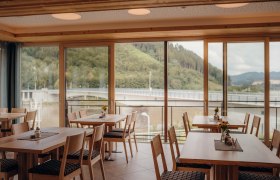 Innenansicht eines Gasthofes mit gro&szlig;en Fenstern und Blick auf eine Br&uuml;cke und bewaldete H&uuml;gel.