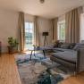 Modernes Wohnzimmer mit grauem Sofa, Holzboden und großem Fenster mit Blick ins Grüne.