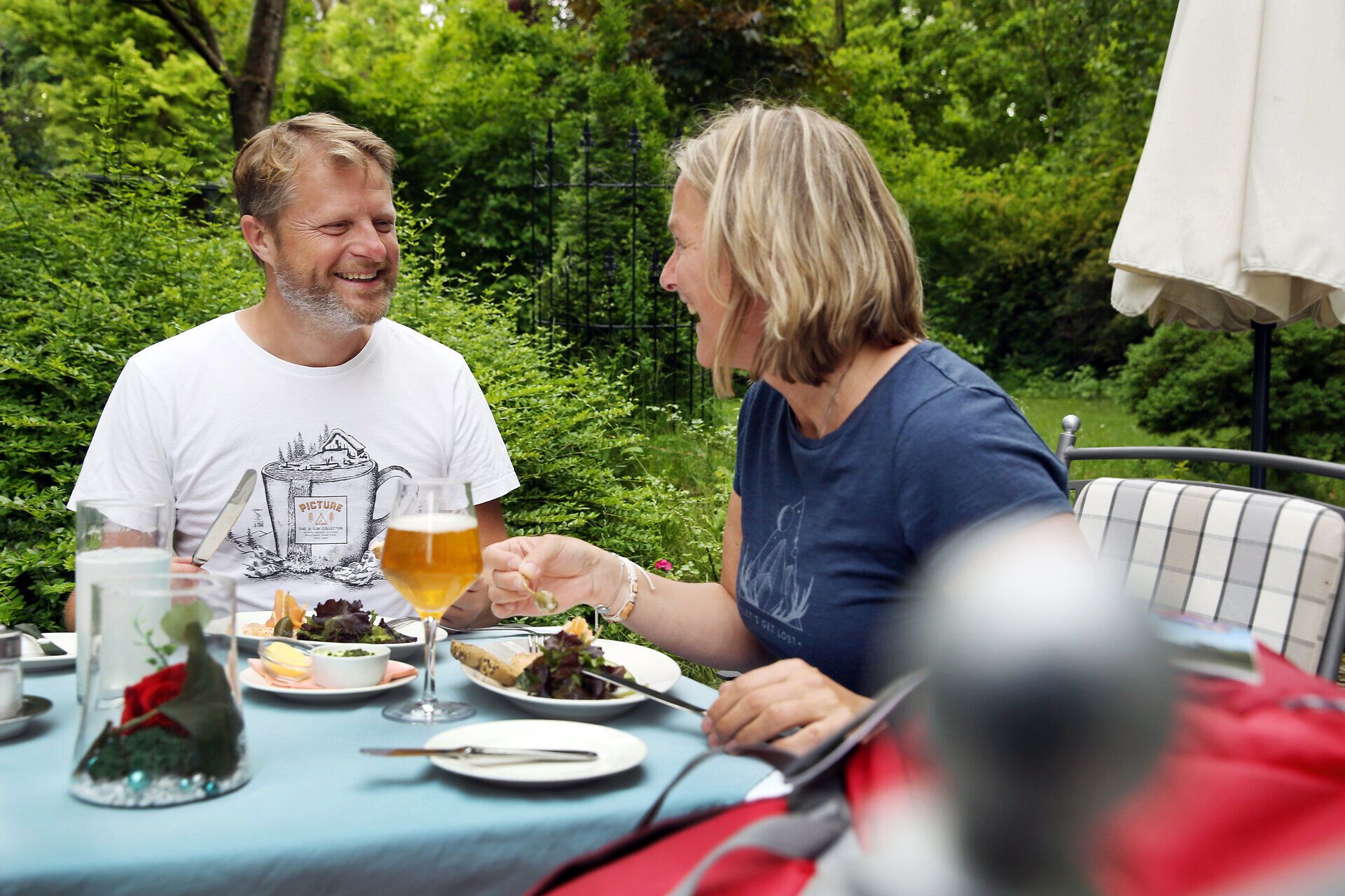 Inmitten einer grünen Oase genießen zwei Wanderer eine entspannte Mahlzeit im Freien. Umgeben von üppigem Grün und dem sanften Plätschern eines Baches, strahlt die Szenerie eine herzliche Gemütlichkeit aus, die zum Verweilen einlädt.
