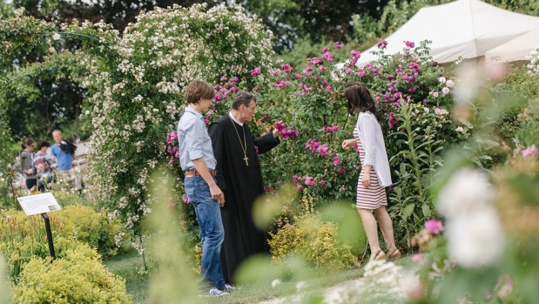 Drei Personen stehen in einem blühenden Garten und unterhalten sich.