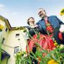 Ein Paar in traditioneller Tracht steht vor einem gelben Geb&auml;ude, umgeben von Blumen, und l&auml;chelt sich an.
