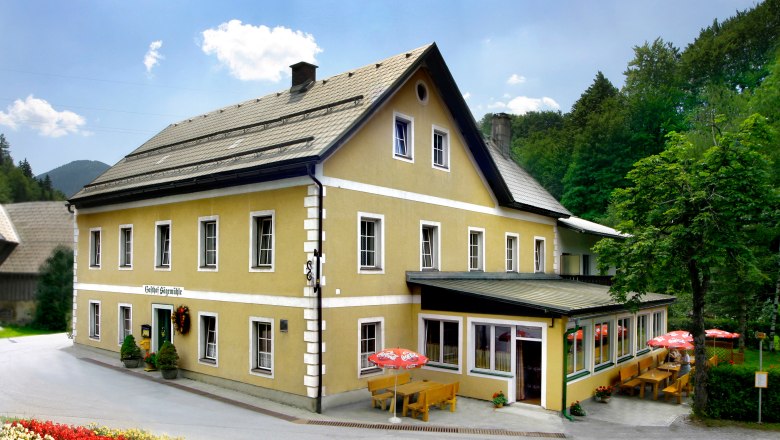 Ein gelbes Gasthaus mit Terrasse und Sonnenschirmen, umgeben von Bäumen.
