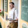 Ein Mann in traditioneller Kleidung lehnt an einer Wand und h&auml;lt ein Tablett mit einem Glas Bier.