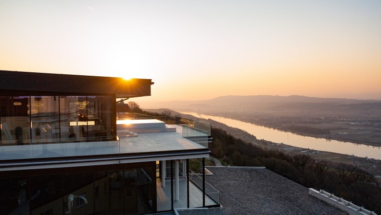 Modernes Gebäude mit Glasfassade bei Sonnenuntergang, Blick auf Fluss und Landschaft.