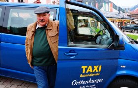 Ein Mann steht neben einem blauen Taxi mit der Aufschrift 'Taxi Botendienste Orttenburger'.