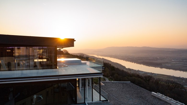 Sonnenuntergang über einem modernen Gebäude mit Infinitypool und Blick auf einen Fluss und Hügel.