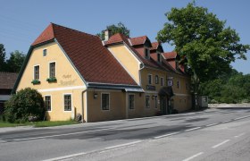Gelbes Gasthaus mit rotem Dach an einer Straße, umgeben von Bäumen.