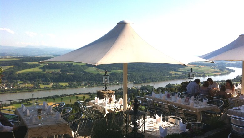 Terrasse mit Tischen und Sonnenschirmen, Blick auf Fluss und Landschaft.