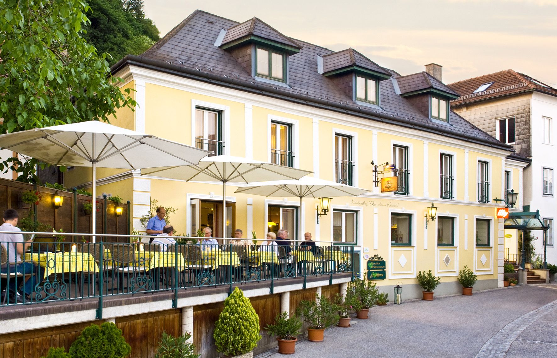 Ein gelber Landgasthof mit Terrasse und Sonnenschirmen, auf der Menschen sitzen.