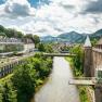 Panorama einer Flusslandschaft mit Schloss und moderner Architektur, umgeben von gr&uuml;nen H&uuml;geln.