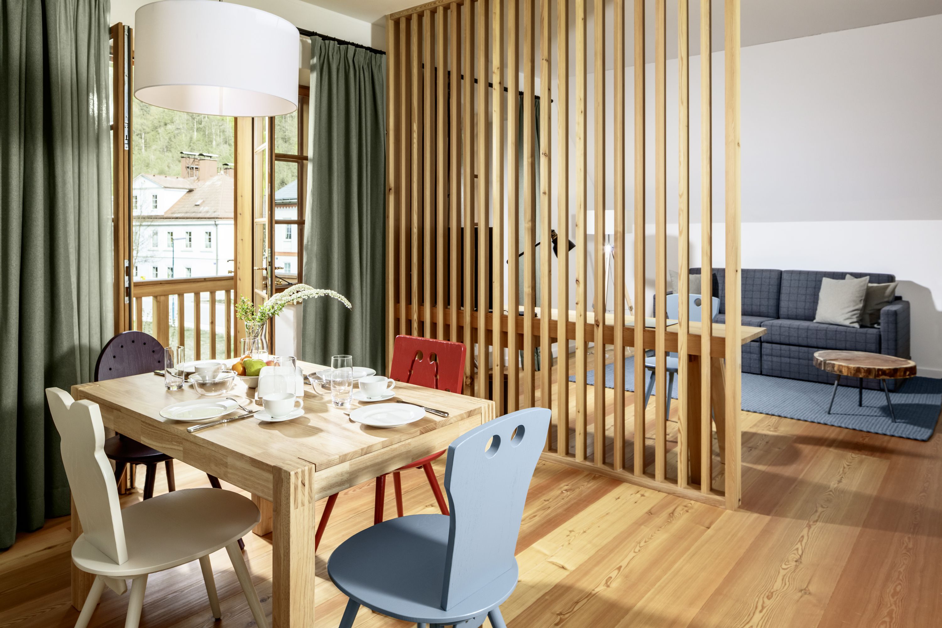 Ein helles Hotelzimmer mit Holzboden, Esstisch und Stühlen, abgetrennt durch eine Holzlamellenwand, mit Blick auf ein Sofa und einen Couchtisch.