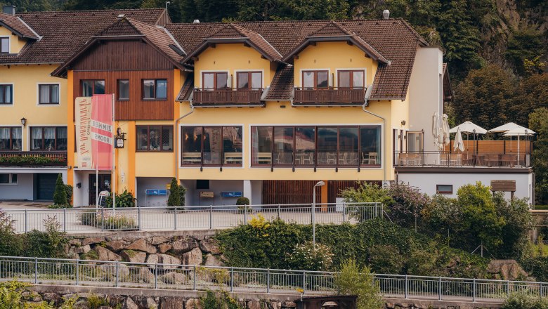 Gelbes Gebäude mit Terrasse und Schildern, umgeben von Bäumen.