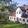 Ein traditionelles Landhaus mit wei&szlig;en W&auml;nden und braunen Fenstern, umgeben von gr&uuml;nen B&auml;umen und einem gepflegten Garten mit einem Picknicktisch.