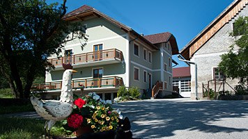 Ein Bauernhof mit einem gr&uuml;nen Haus, Blumen und einer G&auml;nsestatue im Vordergrund.