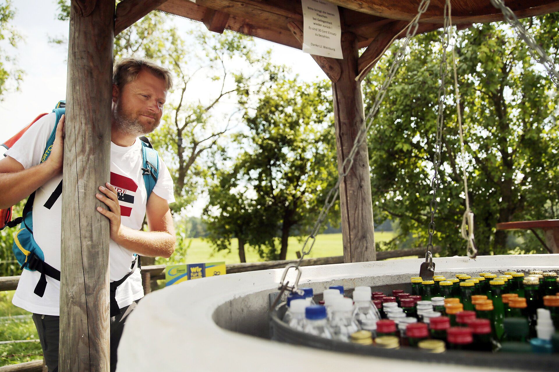 Ein Wanderer genießt eine wohlverdiente Pause an einem malerischen Mostbrunnen, umgeben von üppigem Grün und der sanften Hügellandschaft des Pielachtals. Die frische Luft und die ruhige Atmosphäre laden dazu ein, die Seele baumeln zu lassen und die Schönheit der Natur zu erleben.