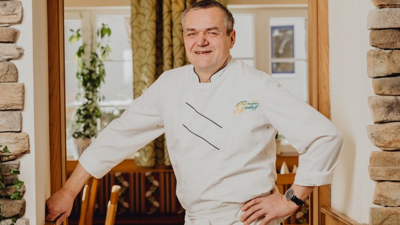 Ein Mann in weißer Kochuniform steht lächelnd in einem Restaurant.