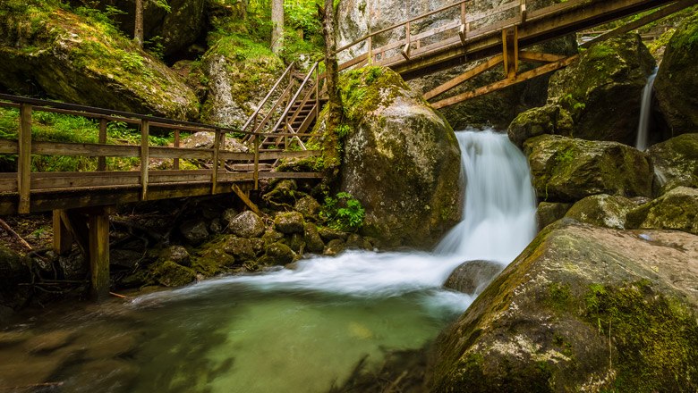 Holzbr&uuml;cken und Wasserfall in einer bewaldeten Schlucht.