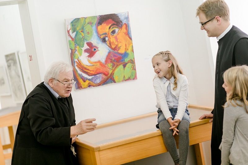Zwei Pater sprechen mit zwei Kindern in einer Galerie vor einem bunten Gemälde.