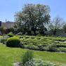Ein gepflegter Garten mit einem Labyrinth aus Hecken, einem großen Baum und einem kleinen Gebäude im Hintergrund bei sonnigem Wetter.