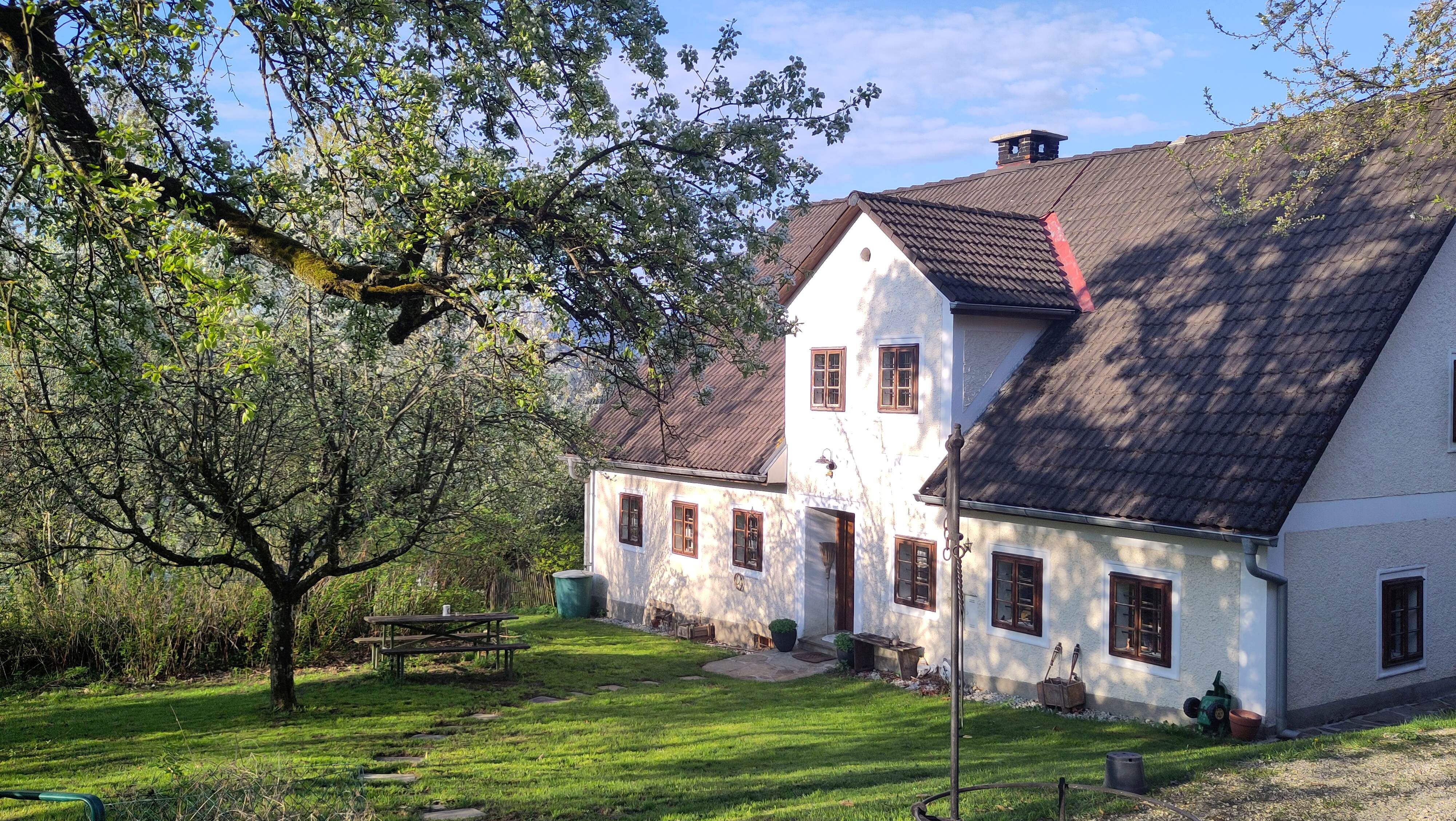 Ein traditionelles Landhaus mit weißen Wänden und braunen Fenstern, umgeben von grünen Bäumen und einem gepflegten Garten mit einem Picknicktisch.