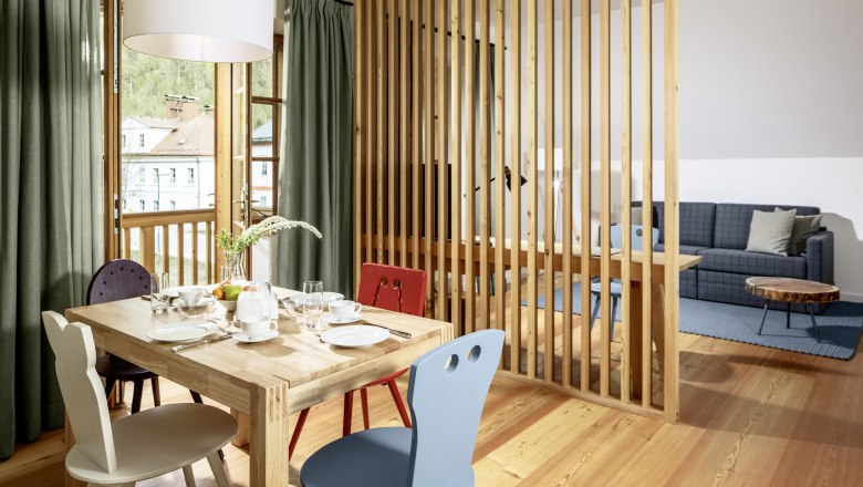 Ein helles Hotelzimmer mit Holzboden, Esstisch und Stühlen, abgetrennt durch eine Holzlamellenwand, mit Blick auf ein Sofa und einen Couchtisch.