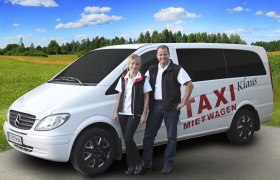 Zwei Personen stehen vor einem weißen Mercedes-Van mit der Aufschrift 'Taxi Mietwagen Klaus' auf einer ländlichen Straße.