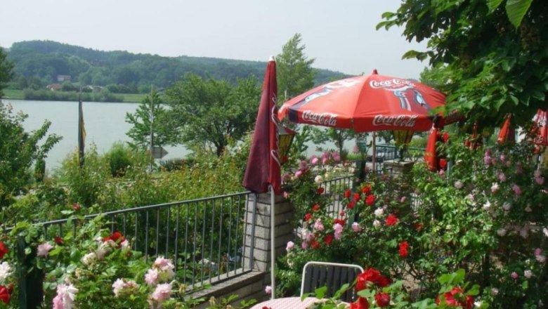 Ein bl&uuml;hender Garten mit roten und rosa Rosen, einem Tisch mit St&uuml;hlen und einem roten Sonnenschirm mit Coca-Cola-Logo, im Hintergrund ein See.