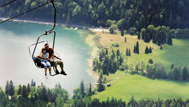 Zwei Personen sitzen in einem Sessellift &uuml;ber einer gr&uuml;nen Landschaft mit einem See im Hintergrund.