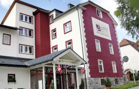 Au&szlig;enansicht eines mehrst&ouml;ckigen Geb&auml;udes mit roter und wei&szlig;er Fassade, beschriftet mit "Zusers Pension".