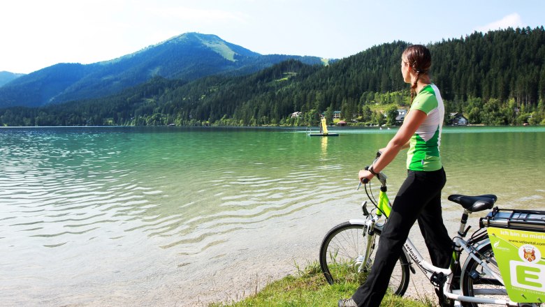 Eine Frau mit Fahrrad steht am Ufer des Erlaufsees und blickt auf das Wasser und die umliegenden Berge.