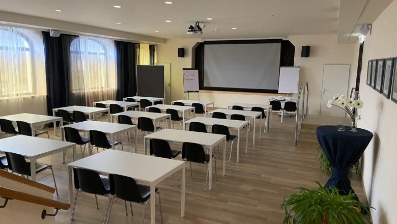 Ein leerer Seminarraum mit weißen Tischen, schwarzen Stühlen und einem großen Bildschirm vorne.