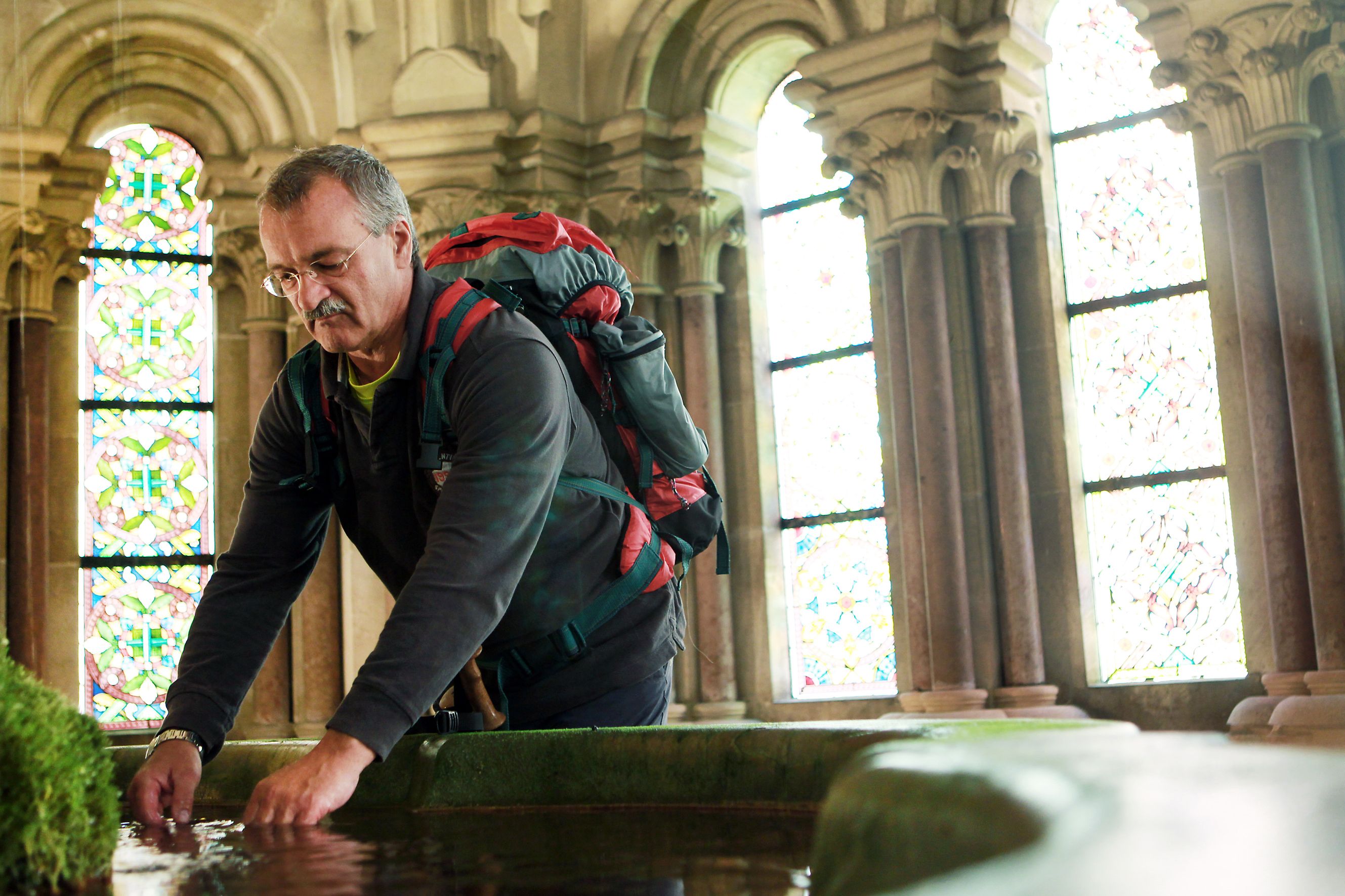 Ein Mann mit Rucksack beugt sich über ein Wasserbecken in einem historischen Gebäude mit bunten Glasfenstern.