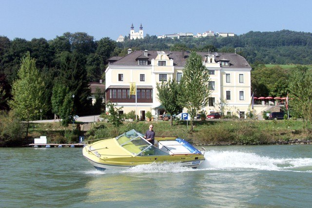 Ein gelbes Motorboot fährt auf einem Fluss vor einem großen, hellen Gebäude mit Hügeln im Hintergrund.