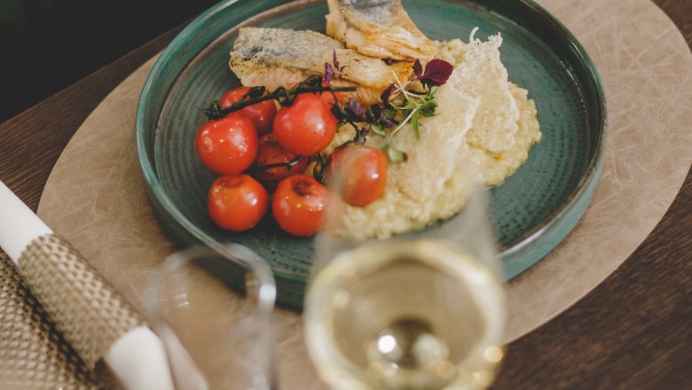 Ein Teller mit gegrilltem Fischfilet, Zitronenrisotto und Kirschtomaten, daneben ein Glas Weißwein.
