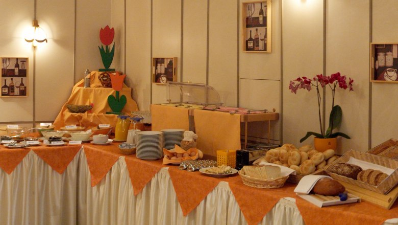 Ein reichhaltiges Frühstücksbuffet mit Brot, Brötchen, Aufstrichen und Getränken auf einem Tisch mit orangefarbenem Tuch.